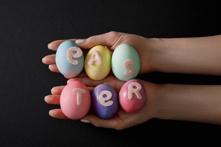 Partial view of woman holding painted chicken eggs with easter lettering on black background