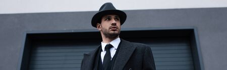 Low angle view of mafioso in black coat and hat with weapon on street, panoramic shot