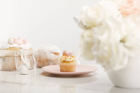 Selective focus of cupcake on plate, decorative rabbits, easter cakes and vase with bouquet on white...