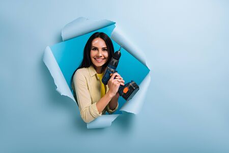 cheerful handywoman with dirty face in paint holding electric drill in torn paper, isolated on blue