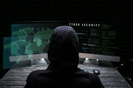 back view of hooded hacker sitting near computer monitors with cyber security lettering on black