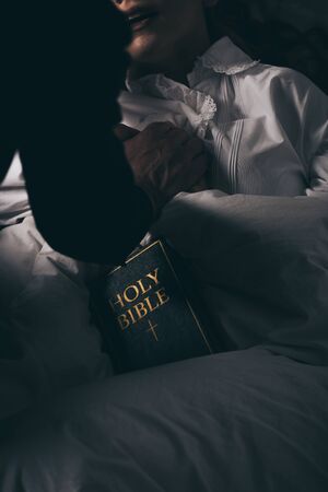cropped view of exorcist with bible holding hands with demonic obsessed girl in bed