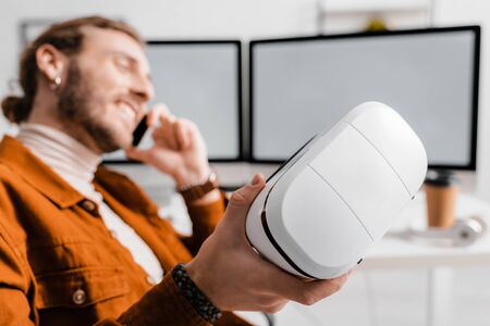 Selective focus of smiling 3d artist talking on smartphone and holding vr headset in office