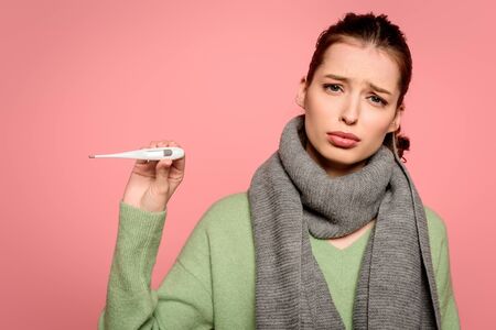 sick, displeased girl in warm showing thermometer isolated on pink