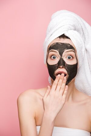 shocked girl with clay mask on face covering mouth with hand and looking at camera isolated on pinkの写真素材