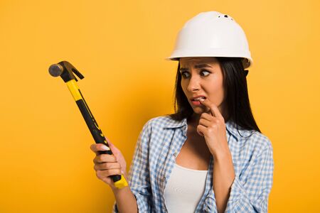frightened female manual worker in helmet holding hammer on yellowの写真素材