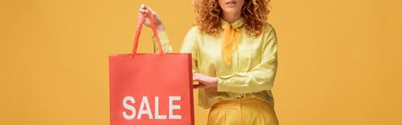 panoramic shot of stylish redhead woman holding shopping bag with sale lettering isolated on yellowの写真素材