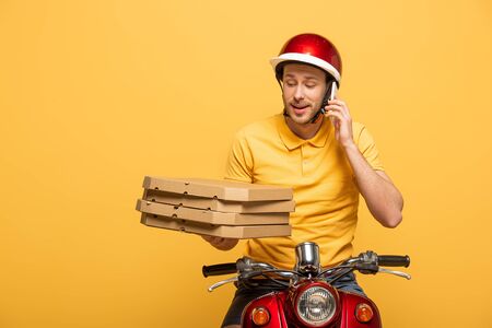 delivery man in yellow uniform riding scooter with pizza boxes and talking on smartphone isolated on yellowの写真素材