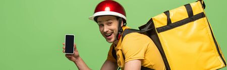 side view of happy delivery man in yellow uniform with backpack showing smartphone with blank screen isolated on green, panoramic shotの写真素材