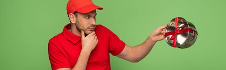 pensive delivery man in red uniform holding pan with bow isolated on green, panoramic shotの写真素材