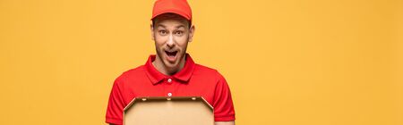 shocked delivery man in red uniform holding pizza box isolated on yellow, panoramic shotの写真素材