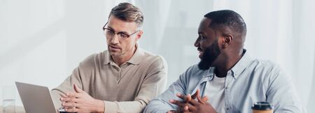 panoramic shot of businessman pointing with hand at laptop and talking with smiling african american...