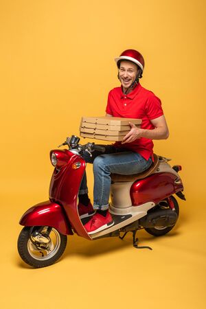 happy delivery man in red uniform holding pizza boxes on scooter on yellow backgroundの写真素材