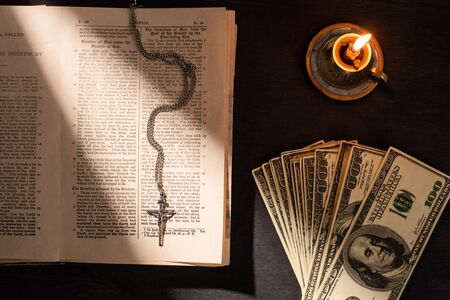 top view of holy bible with money, cross and candle on dark background with sunlightの写真素材