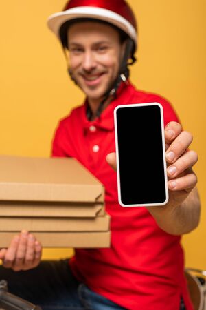 selective focus of happy delivery man in red uniform holding pizza boxes and smartphone with blank screen isolated on yellowの写真素材