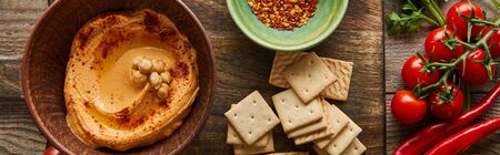 Top view of crackers, bowls with spices and hummus on cutting board with vegetables on wooden background, panoramic shotの写真素材