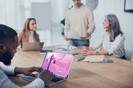 selective focus of african american man using laptop with online shopping website and his colleagues talking on backgroundの写真素材