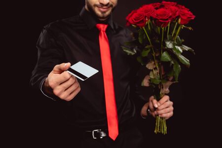 Cropped view of elegant man holding credit card and roses while looking at camera isolated on blackの写真素材