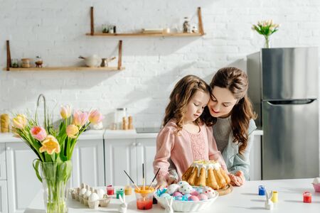 adorable child and happy mother near easter eggs, decorative rabbits, easter bread and tulips