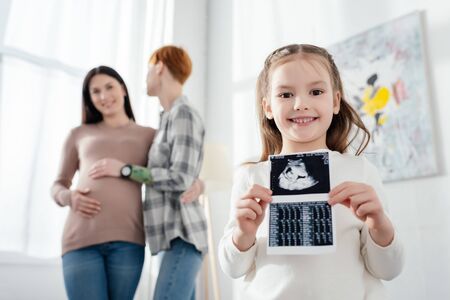 Selective focus of smiling kid holding ultrasound scan of baby near woman embracing pregnant girlfriendの写真素材