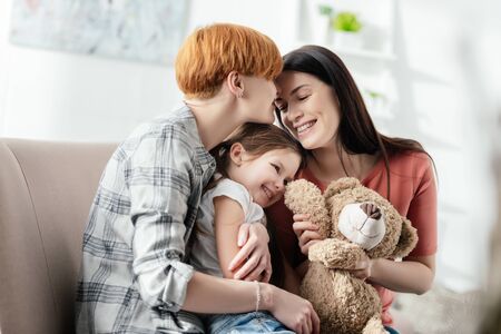 Happy mothers embracing smiling daughter with teddy bear on couchの写真素材
