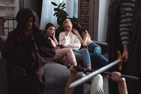 cropped view of men fighting with toy swords near guy in costume of monk while worried girls sitting on sofaの写真素材