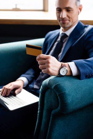 selective focus of smiling businessman in suit holding credit card and using laptopの写真素材