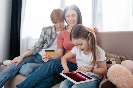 Kid using digital tablet near mothers with credit card on couchの写真素材