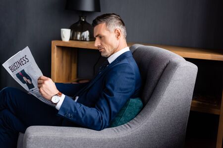 side view of businessman in suit reading newspaper with business lettering and sitting in armchair