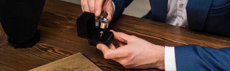 Cropped view of jewelry appraiser examining jewelry ring with magnifying glass at table on grey...