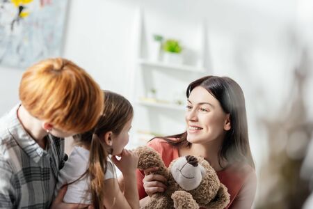 Selective focus of smiling mother holding teddy bear near daughter and parent in living roomの写真素材