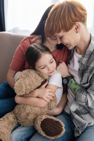 Happy same sex parents hugging daughter with teddy bear on couchの写真素材