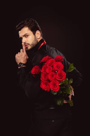 Side view of handsome man holding bouquet of roses and showing secret sign isolated on blackの写真素材