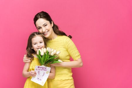 cheerful mother holding greeting card with happy easter lettering and hugging kid with tulips...