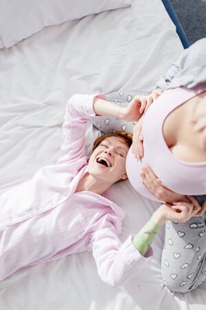 Top view of laughing woman in pajamas touching belly of pregnant girlfriend on bed