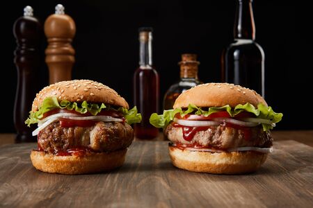 selective focus of two burgers on wooden surface near oil, vinegar and beer bottles, pepper and salt mills isolated on blackの写真素材