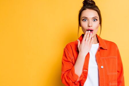 shocked girl looking at camera while covering mouth with hand on yellow backgroundの写真素材