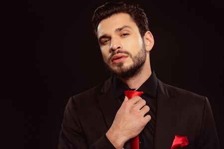 Elegant man looking at camera and adjusting red tie isolated on blackの写真素材
