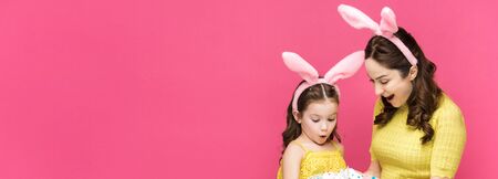 panoramic shot of surprised mother and daughter in bunny ears looking at easter eggs isolated on...