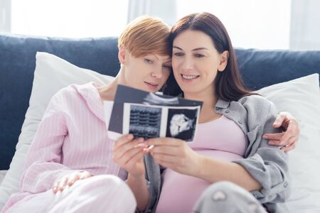 Selective focus of smiling same sex couple in pajamas looking at ultrasound scans of baby on bedの写真素材