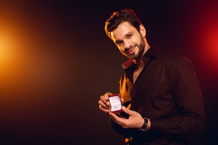 Handsome man holding gift box with jewelry ring and smiling at camera on black background with lightingの写真素材