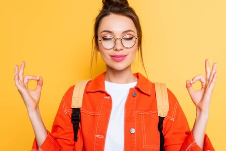 smiling student in eyeglasses standing in meditation pose with closed eyes isolated on yellowの写真素材