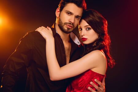 Beautiful woman in red dress looking at camera while hugging handsome man on black background with lightingの写真素材