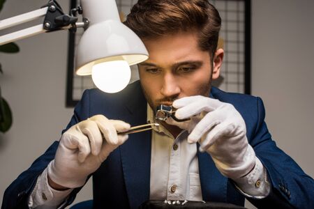 Handsome jewelry appraiser working with gemstone in tweezers and magnifying glass at table in...
