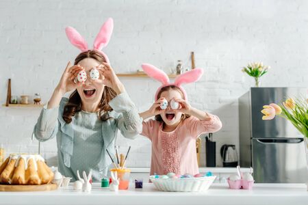 excited mother and daughter in bunny ears covering eyes with painted chicken eggs near tulips and easter breadの写真素材