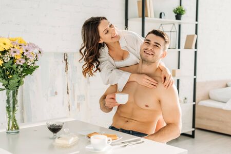 cheerful sexy girl hugging happy shirtless boyfriend drinking coffee while having breakfastの写真素材
