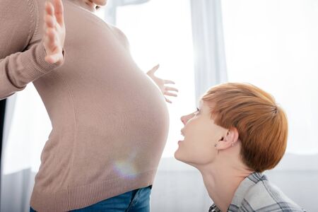 Side view of woman looking at belly of pregnant girlfriend at homeの写真素材