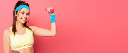 attractive sportswoman training with dumbbell while smiling at camera on pink backgroundの写真素材