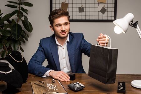 Handsome jewelry appraiser holding paper bag near earrings and necklaces on tableの写真素材