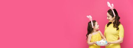 panoramic shot of happy mother and daughter in bunny ears holding easter eggs isolated on pinkの写真素材
